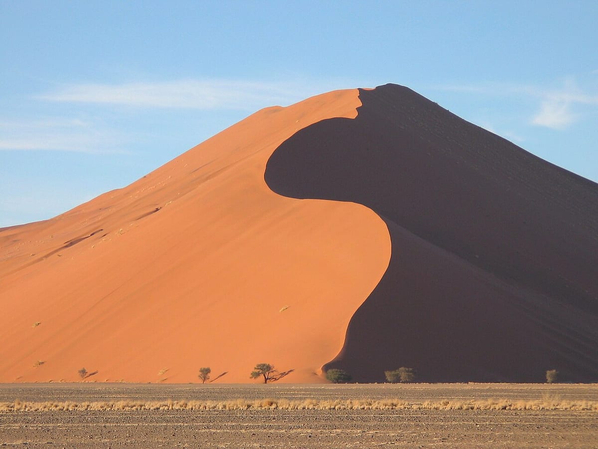Eine rote Sanddüne mit scharfer Schattenkante, davor eine trockene Ebene