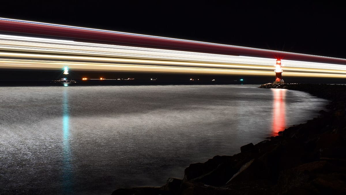 Langzeitbelichtung an einer Küste: das Wasser verwischt zu einer glatten Fläche, Lichter ziehen Spuren